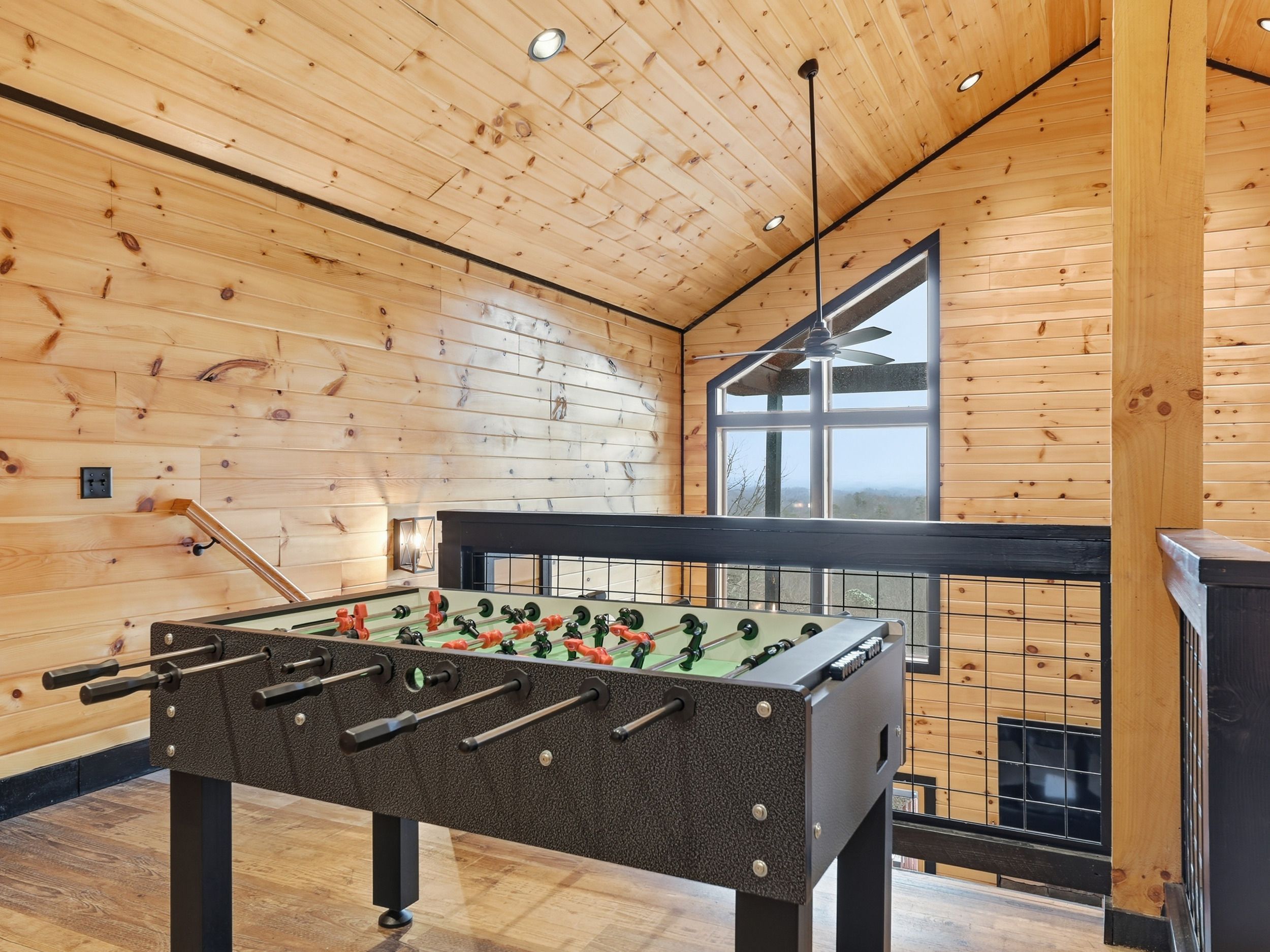 Una habitación con piso de madera, mesa de fútbol, ventilador de techo y un ventanal con vistas.