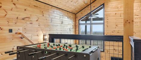 Una habitación con piso de madera, mesa de fútbol, ventilador de techo y un ventanal con vistas.