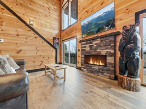 A cozy living room with wooden walls, a stone fireplace, a leather sofa, and a large window with a mountain view.
