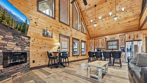A spacious log cabin interior with a stone fireplace, wooden dining area, kitchen, and a large window offering a mountain view.