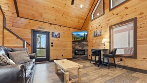 A cozy cabin interior with a leather sofa, a wooden coffee table, a stone fireplace, and a dining area with two chairs.