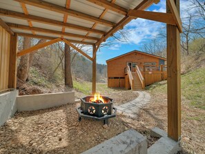 A wooden cabin with a fire pit under a covered patio, surrounded by a natural landscape. - Luxury Cabin w/Huge Game Room, Fire Pit & Hot Tub (Pigeon Forge)