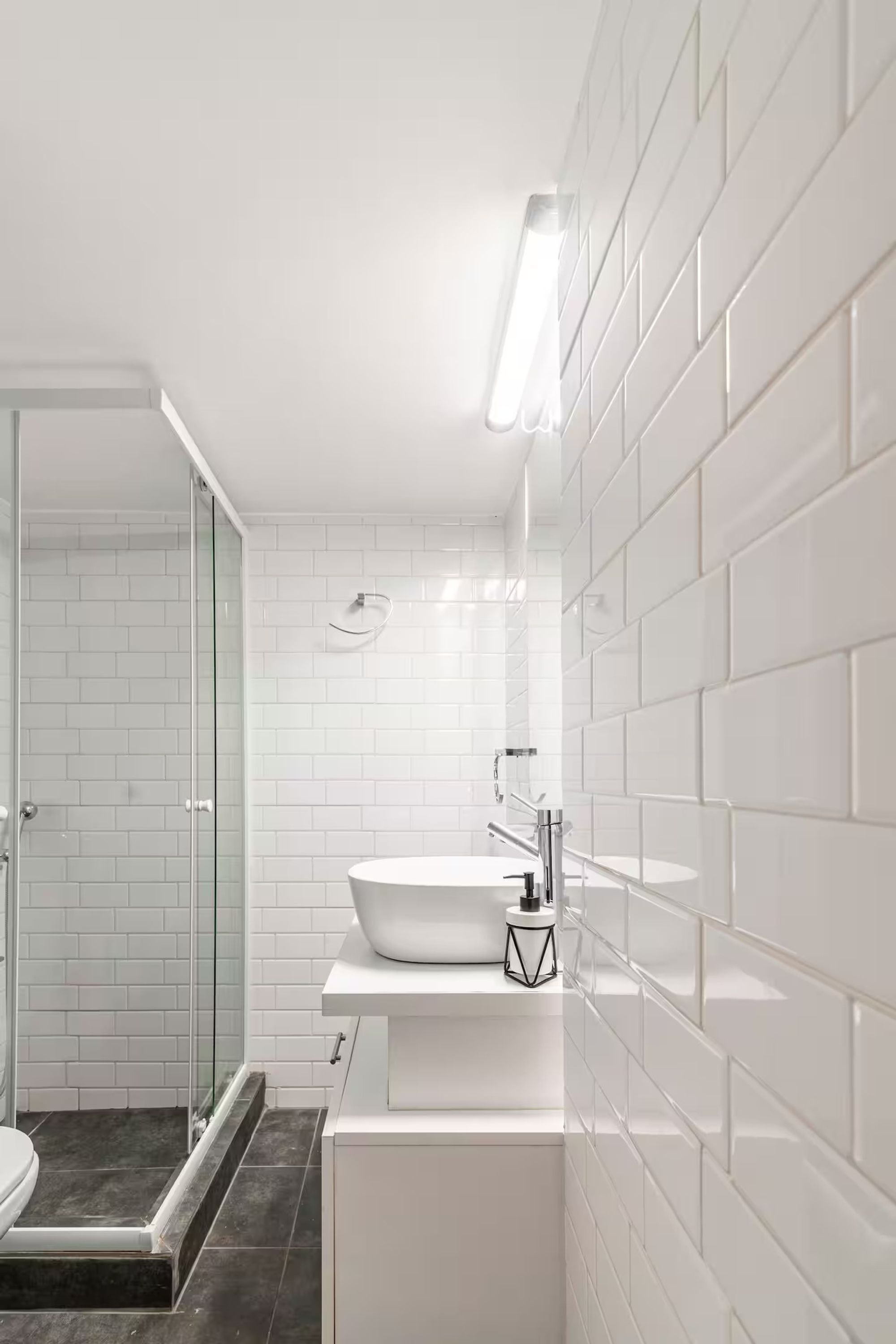 Um banheiro moderno com azulejos brancos tipo metrô, box de vidro e pia branca com porta-sabonete preto geométrico.