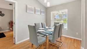 A dining area with a glass table, grey upholstered chairs, and a window with blinds. - Waterview 3 Bed In Sunridge Woods 3 Bedroom Villa (Davenport)