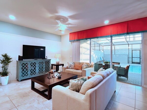 A living room with a sofa, television, and a view of a pool through large windows. - Fabulous 5bd Family Townhome With Splash Pool-726g 5 Bedroom Townhouse (Davenport)