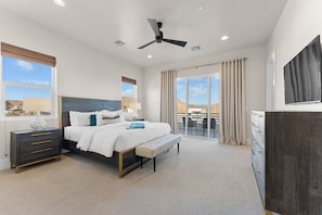 Une chambre à coucher comprenant un lit, des tables de chevet, un ventilateur de plafond, une grande fenêtre et une vue sur l’extérieur.