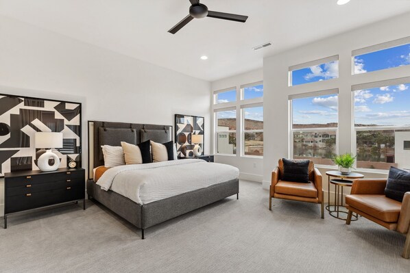 A modern bedroom with a large bed, a nightstand, a chair, and a view of the landscape through large windows. - Villa #36 At Bloomington Country Club 4 Bedroom Townhouse (St. George)
