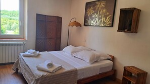 Una cama bien tendida con ropa y toallas blancas, un separador tejido, una lámpara de pie, un cuadro botánico enmarco y un pequeño taburete de madera.