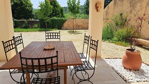 Un patio al aire libre con una mesa de madera y cuatro sillas, rodeado de grava y follaje.