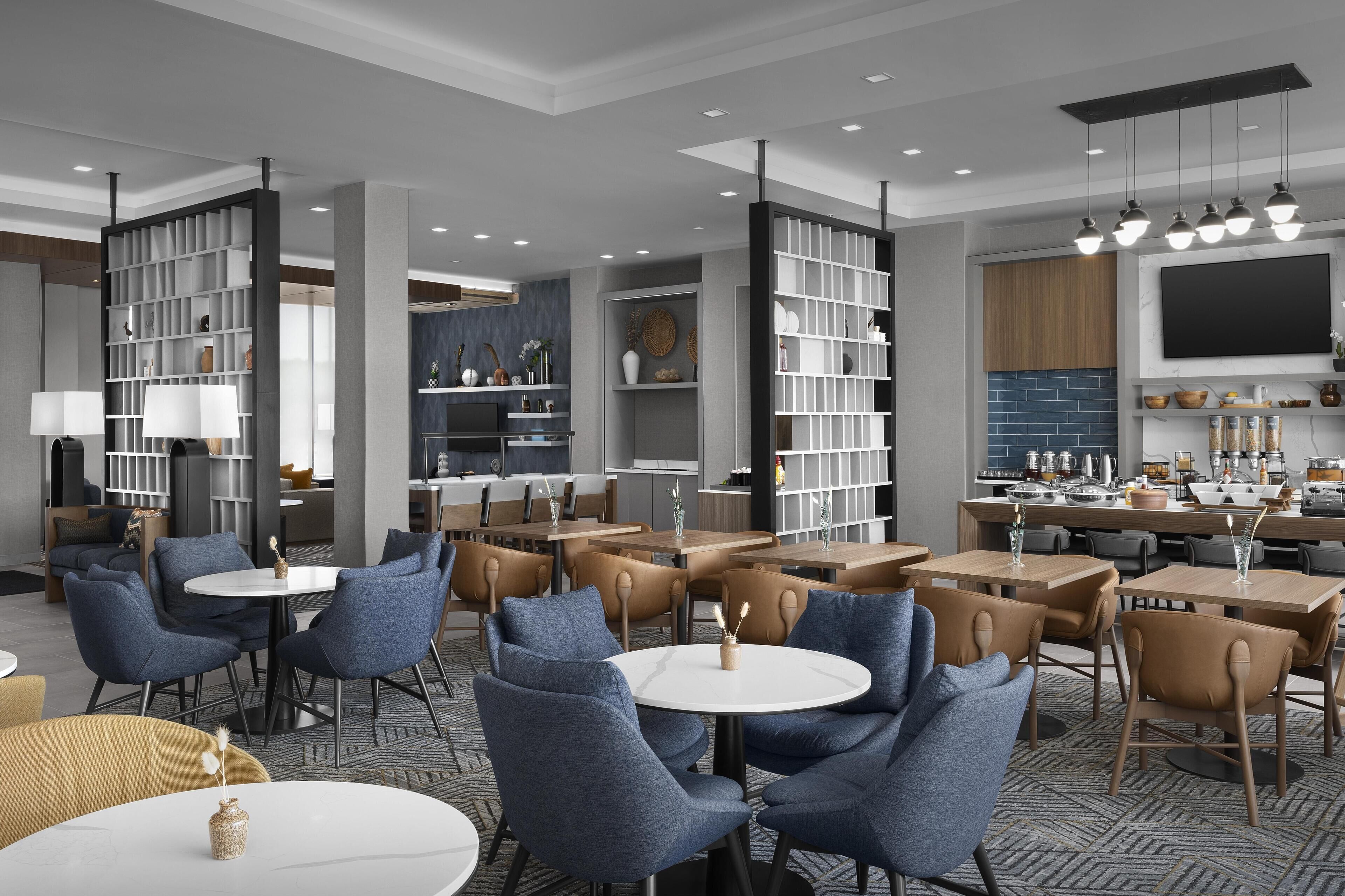 A modern dining area with round tables and blue chairs, surrounded by bookshelves and a kitchen area in the background.