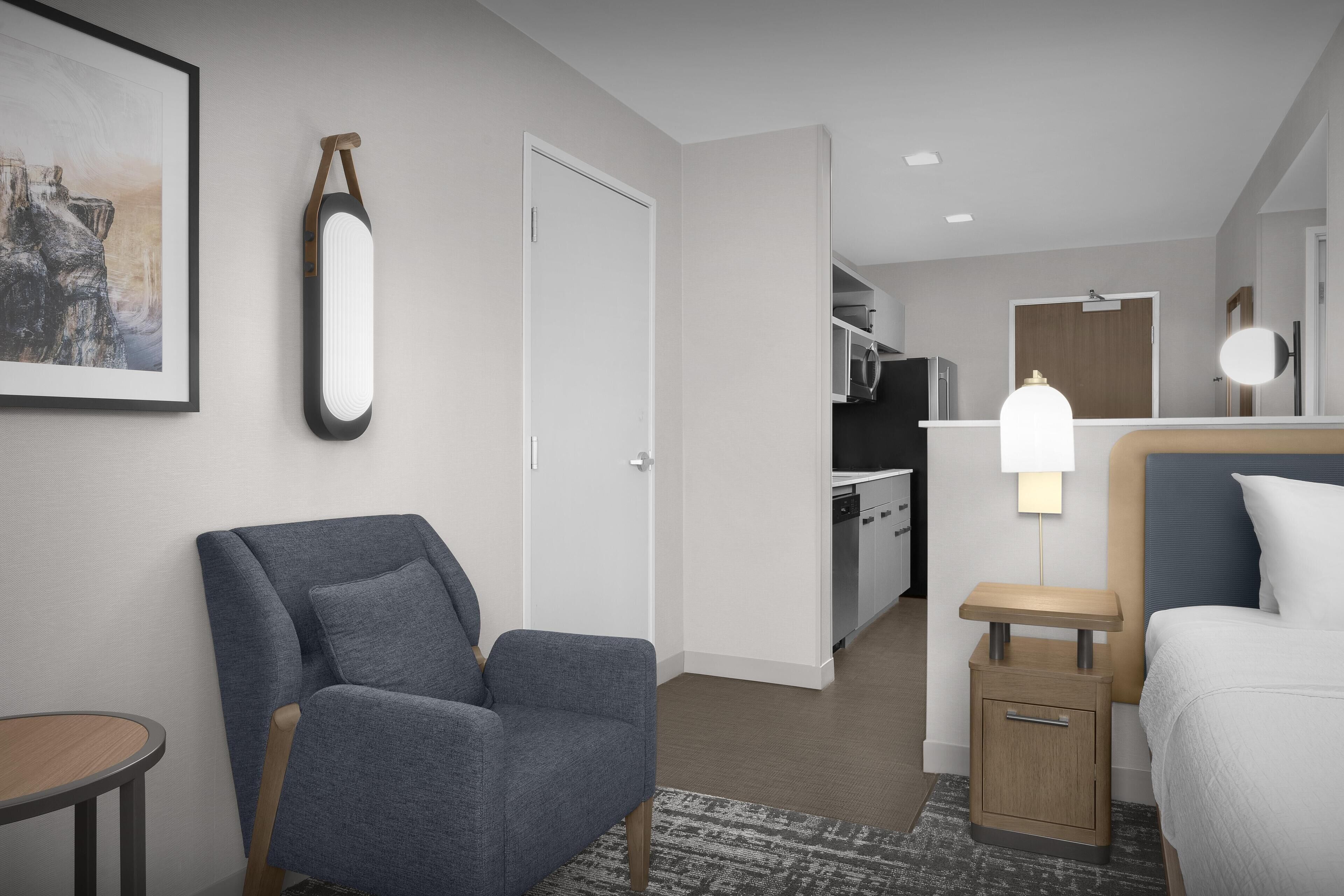 A hotel room with a grey armchair, a bedside table with a lamp, a bed, a small table, and a framed picture on the wall.