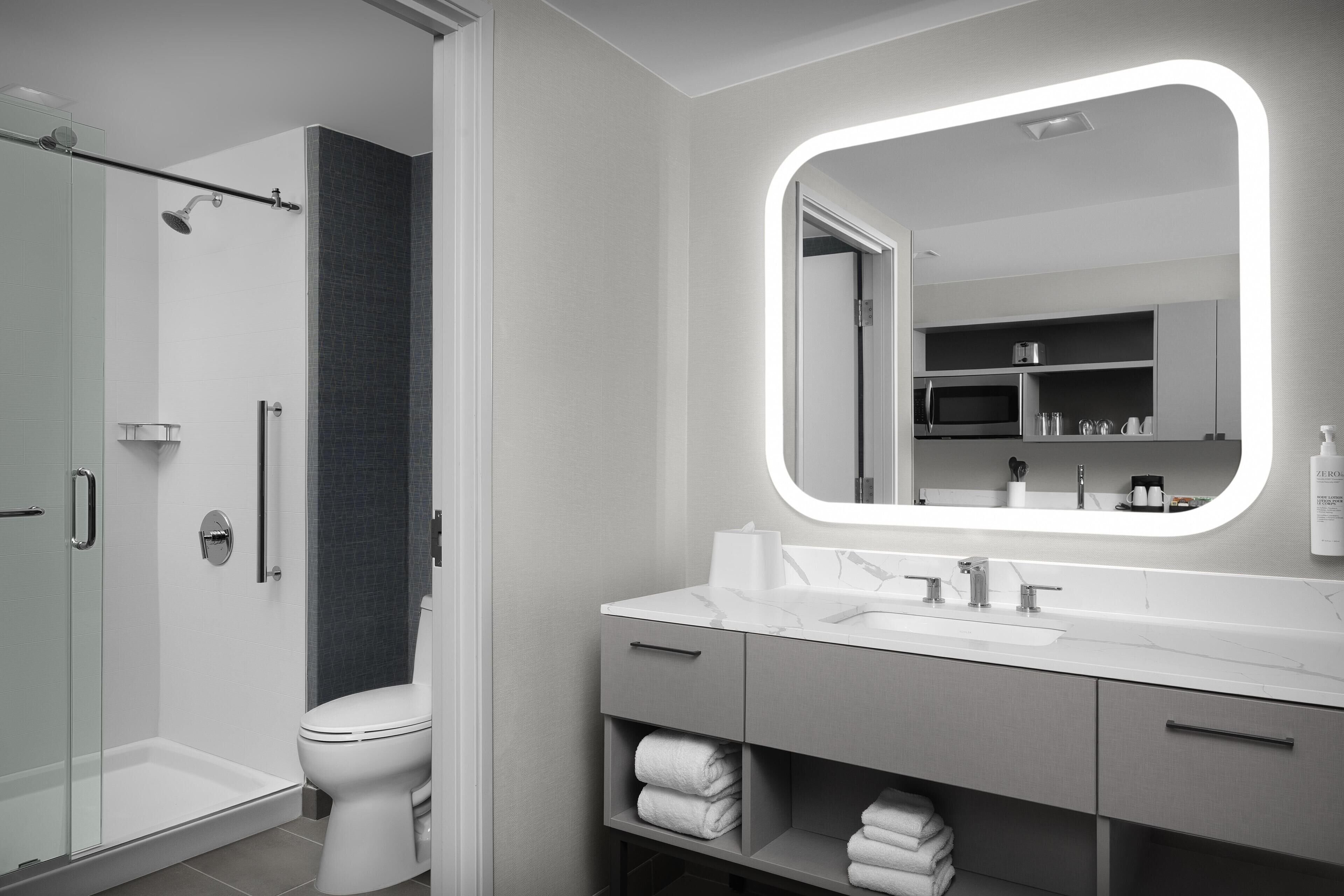 Un baño moderno con cabina de ducha de vidrio, inodoro blanco, tocador con un espejo grande y toallas ordenadas en estantes.