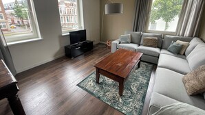A modern living room with a grey sofa, a wooden coffee table, a TV on a black stand, and a view of buildings through large windows. - Topsleep Apartments 26-1 (Arnhem)