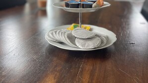 A three-tiered dessert stand with various sweets on a wooden table in a room with a window and a chair in the background. - Topsleep Apartments 26-1 (Arnhem)