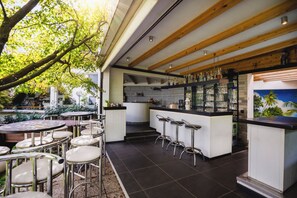 Um café ao ar livre com mesas redondas e banquetas, uma área de cozinha com balcão e um mural na parede.