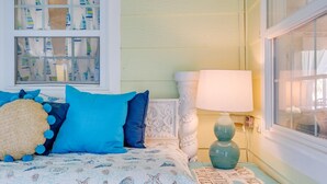 Ein Schlafzimmer mit einem Bett, das mit einer Betttdecke im Seestil ausgestattet ist, einem Fenster mit gestreiften Vorhängen und einem Nachttisch mit Lampe und Dekoration.