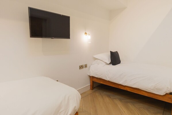 Una habitación pequeña y limpia con dos camas individuales, un televisor de pantalla plana montado en la pared y una lámpara sencilla.