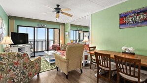 A living room with a television, sofa, armchair, dining table, and chairs. There is a ceiling fan, a window with a view, and a wall sign.
