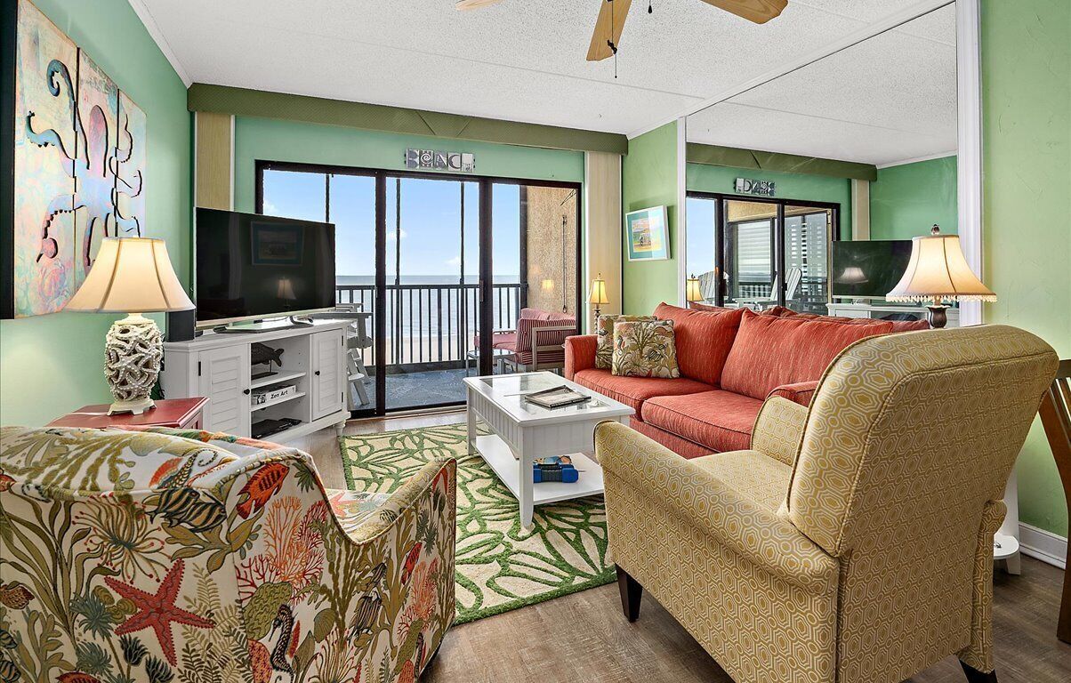 A living room with a red sofa, patterned armchairs, a coffee table, and a TV. There is a balcony with a view.