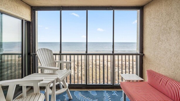 A balcony with a view of the beach, white chairs, a red-striped sofa, and a blue patterned rug.