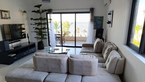En stue med en beige sofa, et sofabord i glas, et fjernsyn og en potteplante.
