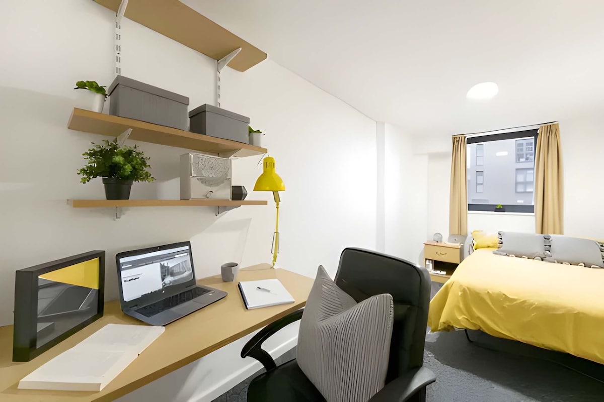 A hotel room with a desk, laptop, chair, bed, bedside table, lamp, and a window with curtains.