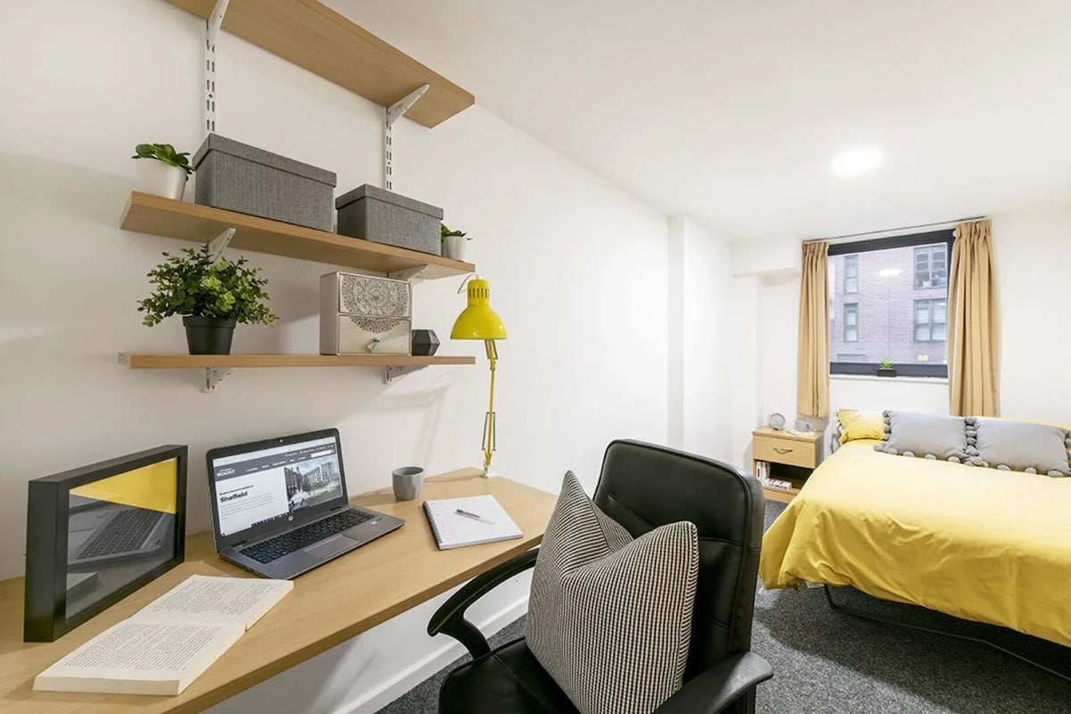 A hotel room with a desk, two laptops, a chair, a bed with a yellow bedspread, a window with curtains, and shelves with decorative items.