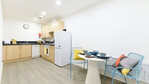A modern kitchen with wooden cabinets, a white refrigerator, and a dining area with blue chairs and a white table. - For Students Only Central Pl Sheffield (Sheffield)