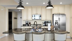 A kitchen with three bar stools, a central island, and a stainless steel refrigerator.