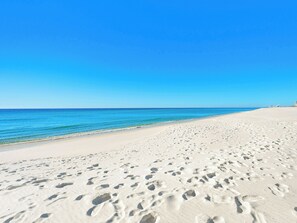 Een breed zandstrand met voetafdrukken die naar het water leiden, een heldere blauwe lucht en een rustige zee.