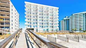 Un muelle de madera que conduce a un hotel frente a la playa, con varios balcones y un cielo azul despejado.