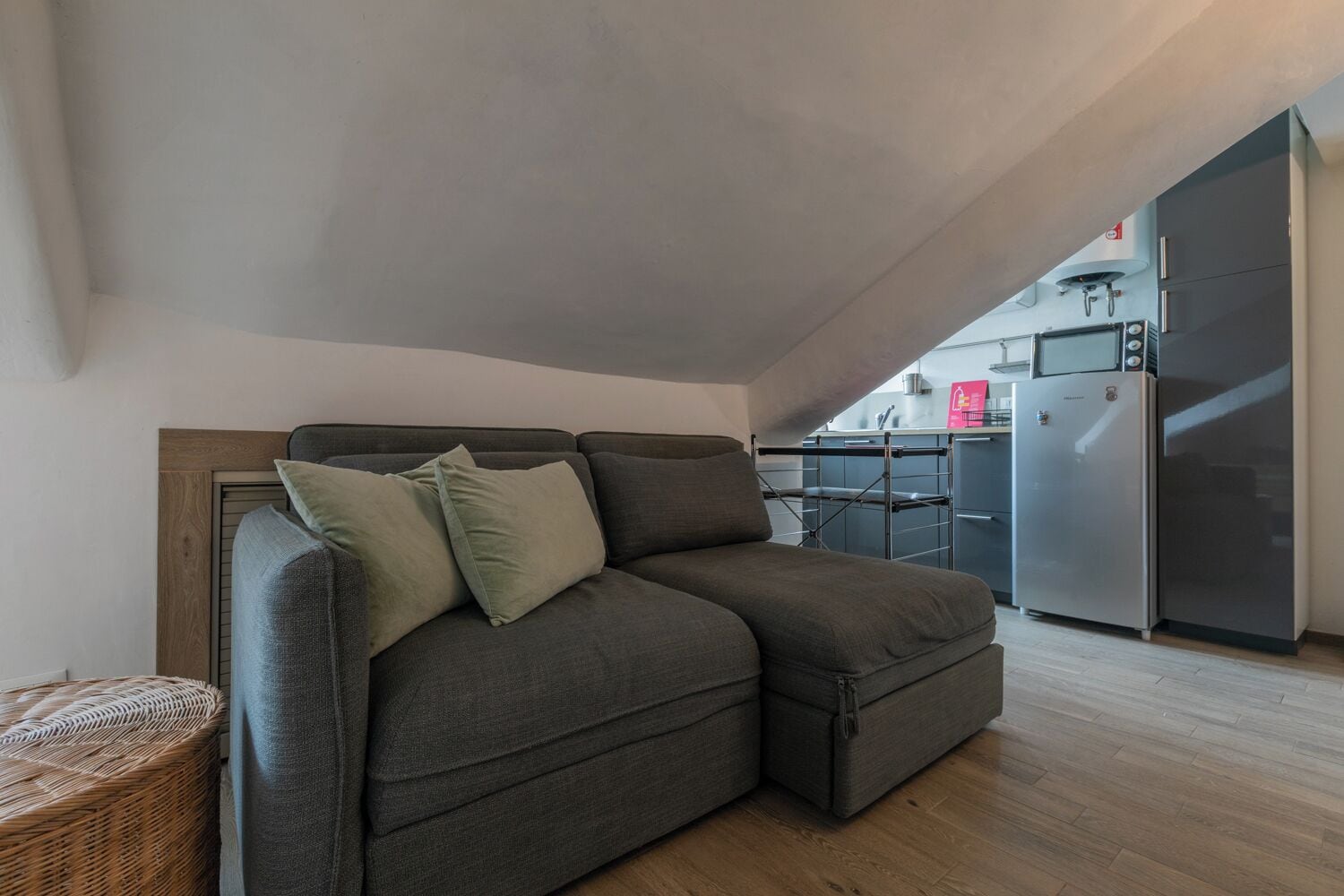 A modern living room with a grey sectional sofa, a small wicker side table, and a kitchen area with a stainless steel refrigerator and microwave.
