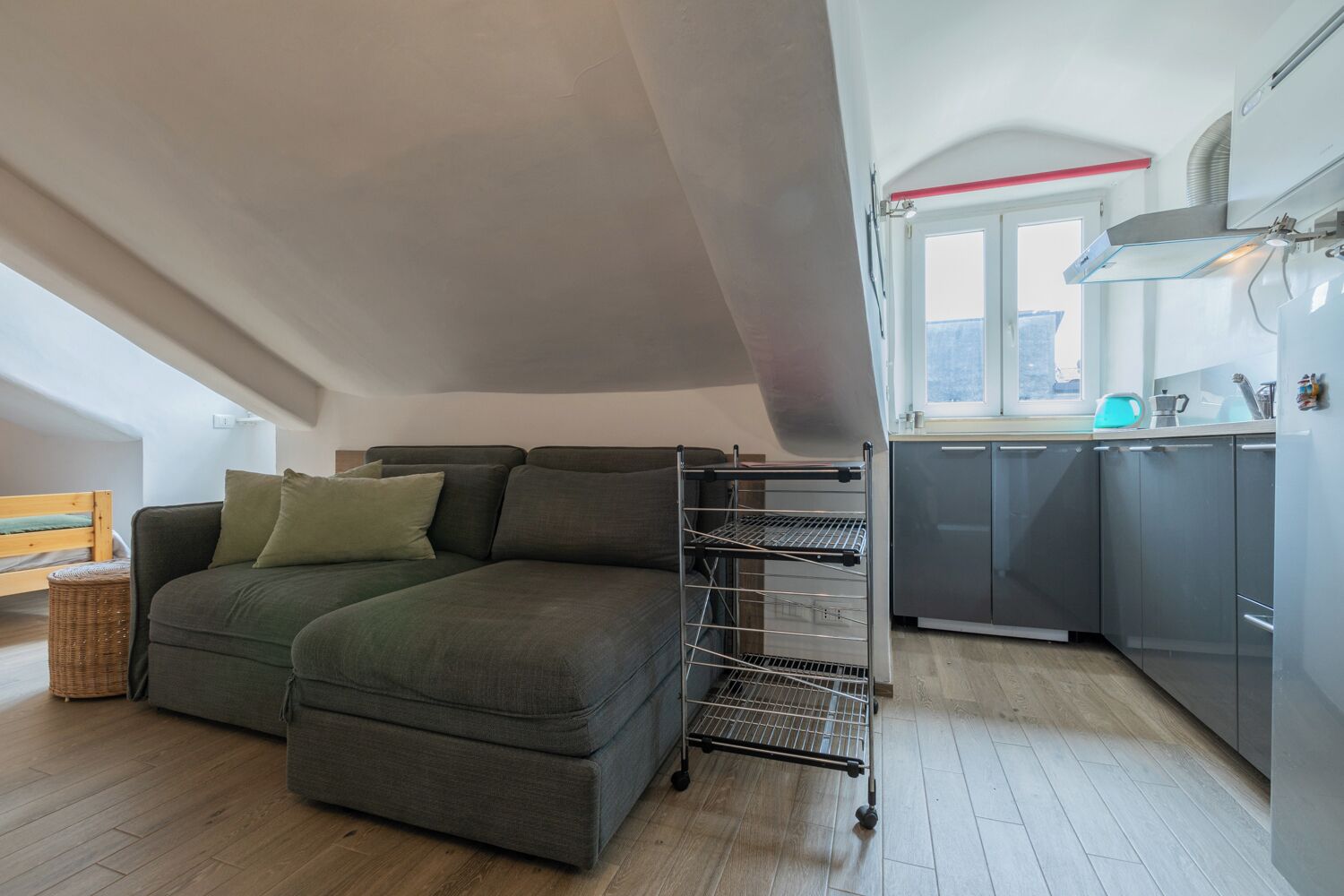 Un espace de vie compact comprenant un canapé, une étagère en métal, un coin cuisine avec des meubles gris et une fenêtre laissant entrer la lumière naturelle.