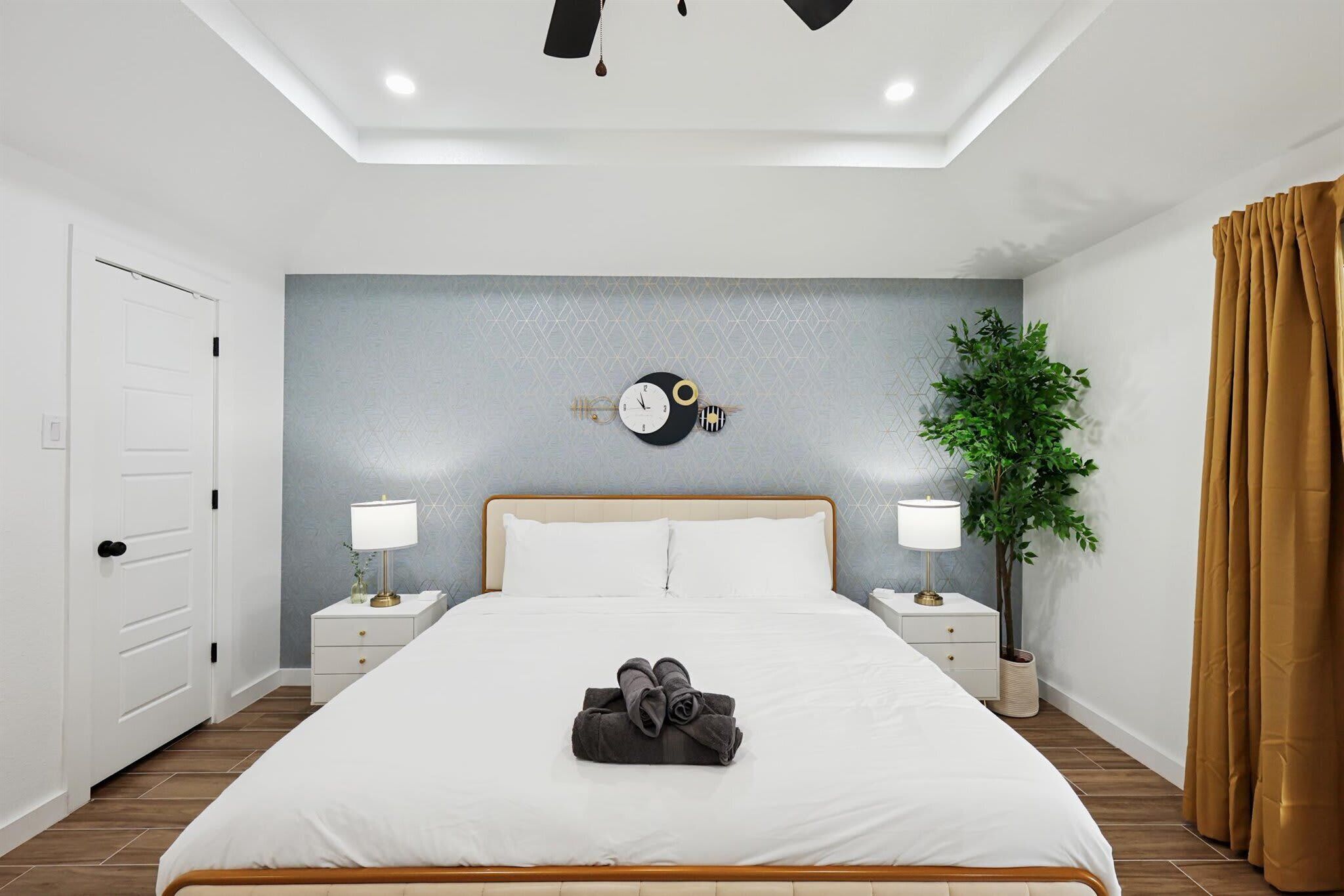A modern hotel room with a large bed, two bedside tables with lamps, a wall clock, a potted plant, and a door.
