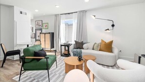 Ein modernes Wohnzimmer mit einem grünen Sofa, einem weißen Sessel und einem grauen Sofa mit gelben Kissen. Zwei Holztische und eine Stehlampe sind ebenfalls vorhanden.