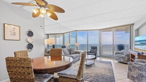 A spacious living area with a large dining table, wicker chairs, a sofa, and a TV. The room has large windows offering a view of the ocean.