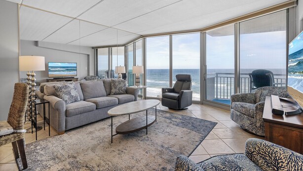 A modern living room with a grey sofa, armchairs, a coffee table, and a TV. Large windows offer a view of the ocean.