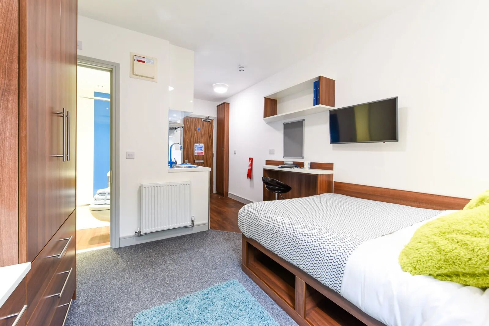 A modern hotel room with a bed, a desk, a chair, a TV, and a bathroom visible through an open door.