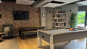Un salon moderne doté d’une table de billard, d’un fauteuil confortable, d’une bibliothèque et d’une grande fenêtre offrant une vue sur la verdure.