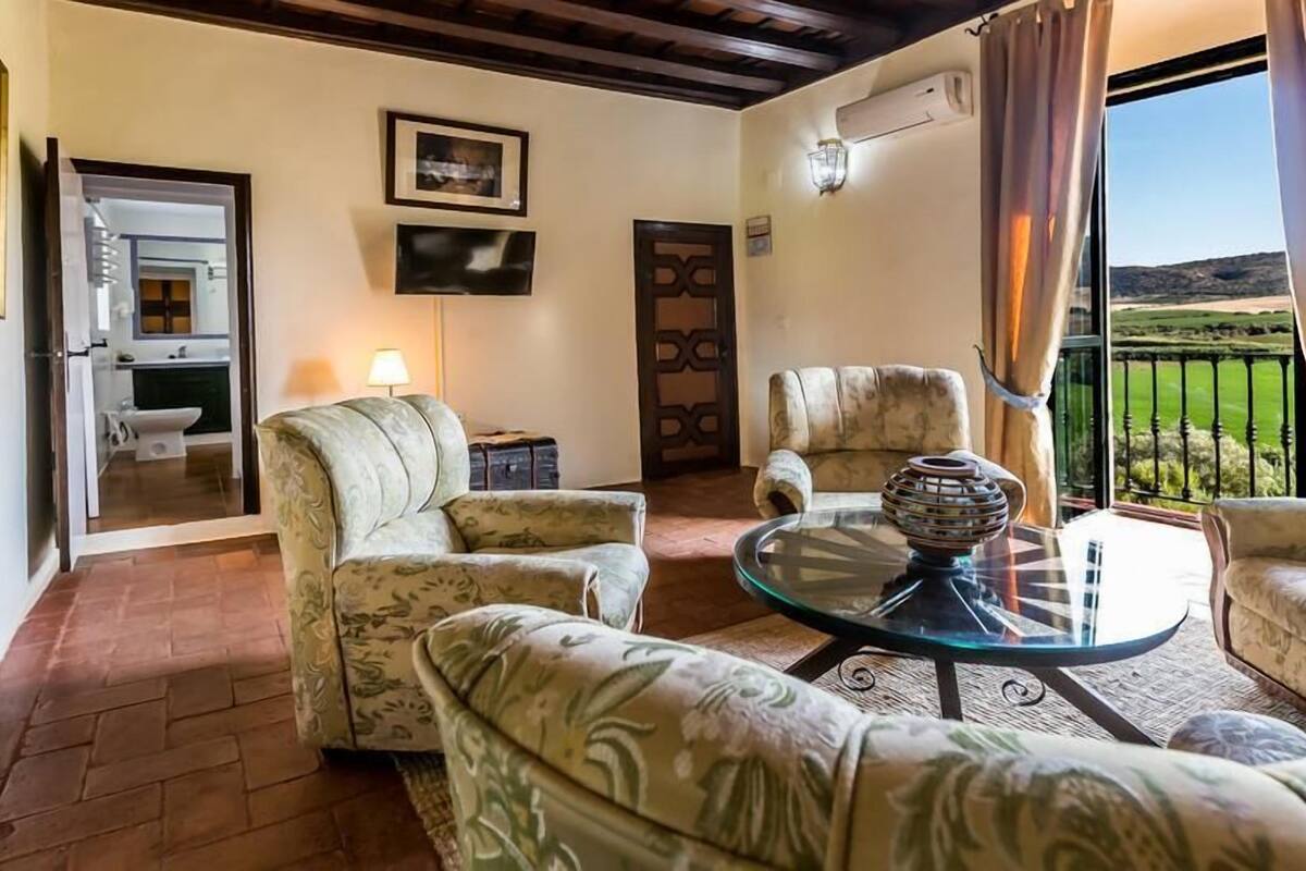 Sala de estar con una mesa de centro de vidrio, dos sillones estampados, un sofá y vistas a un paisaje verde a través de un balcón.