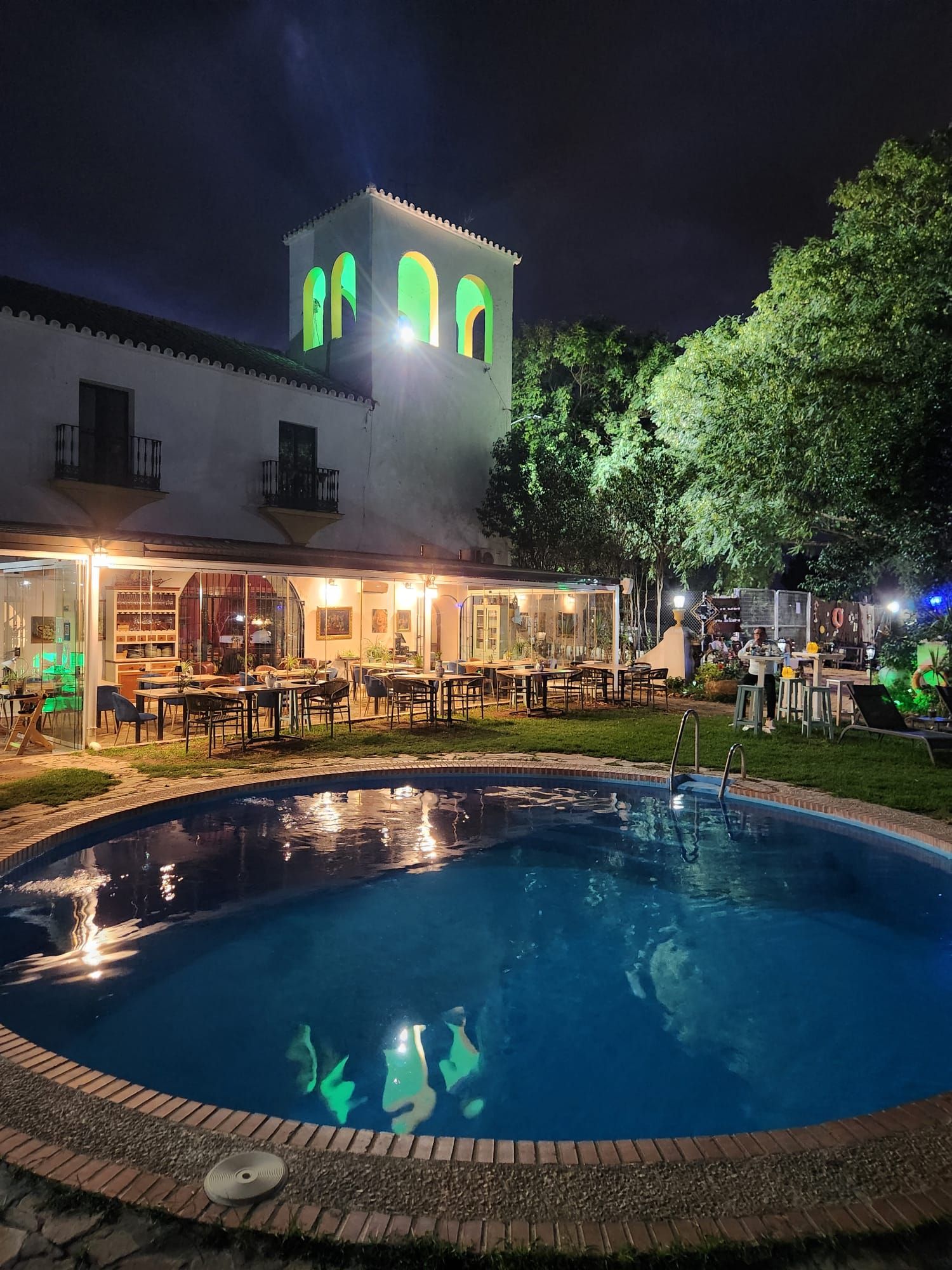 Zona de piscina con un edificio que tiene ventanas arqueadas y una torre, rodeada de vegetación.