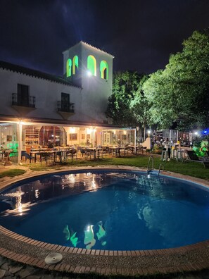Zona de piscina con un edificio que tiene ventanas arqueadas y una torre, rodeada de vegetación.