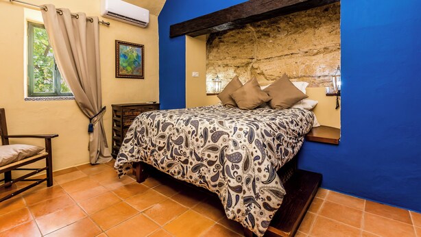 Un dormitorio con cama, una silla, una ventana con cortinas y una pared de piedra.