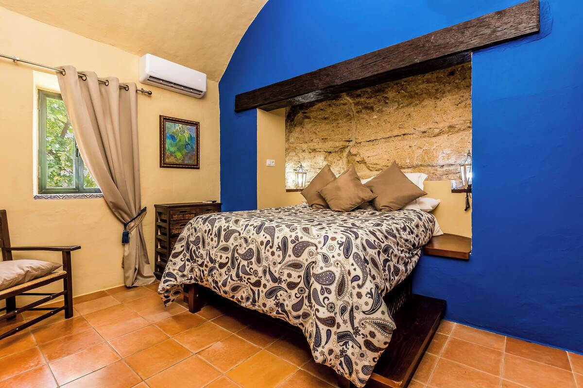 Un dormitorio con cama, una silla, una ventana con cortinas y una pared de piedra.