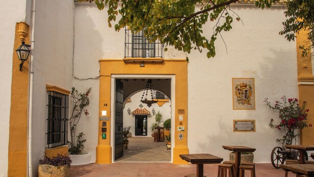 Uma área de pátio ao ar livre com mesas e bancos de madeira, piso ladrilhado e um portal que dá para um espaço interno.