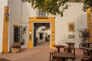Un patio al aire libre con mesas y bancos de madera, piso de baldosas y una puerta de acceso a un espacio interior.