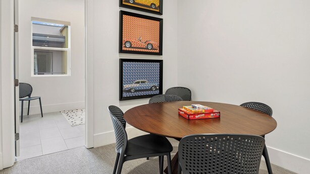 A dining area with a round wooden table, black perforated chairs, and three framed automotive-themed artworks on the wall. - Villa #36 at Bloomington Country Club: Pool, Golf, Restaurant (St. George)