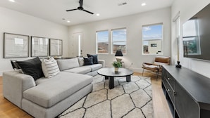 A modern living room with a sectional sofa, a round coffee table, and a TV on a cabinet. There are large windows and a ceiling fan. - Villa #36 at Bloomington Country Club: Pool, Golf, Restaurant (St. George)
