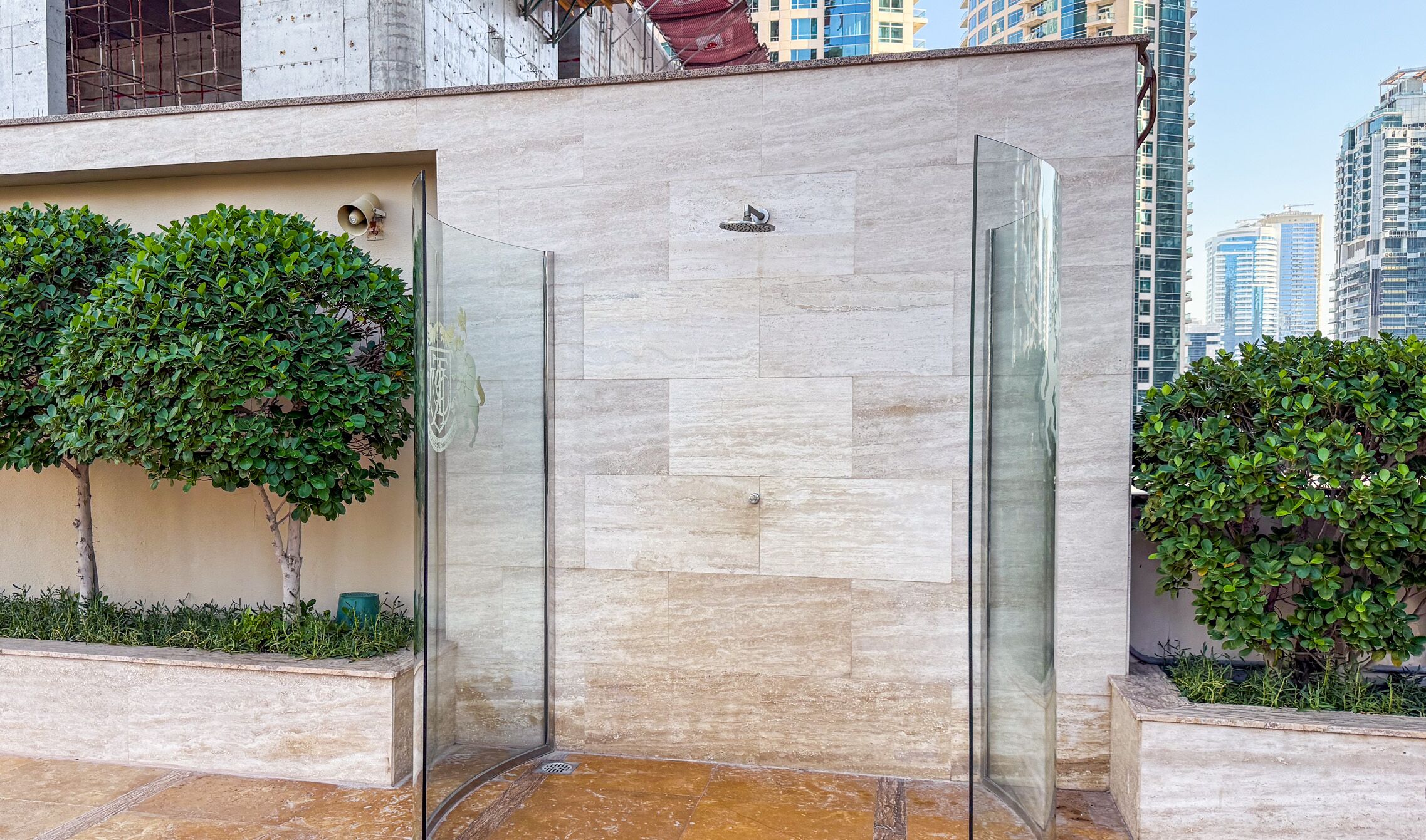 A modern glass-enclosed shower area with a tiled floor, surrounded by greenery and a cityscape view.