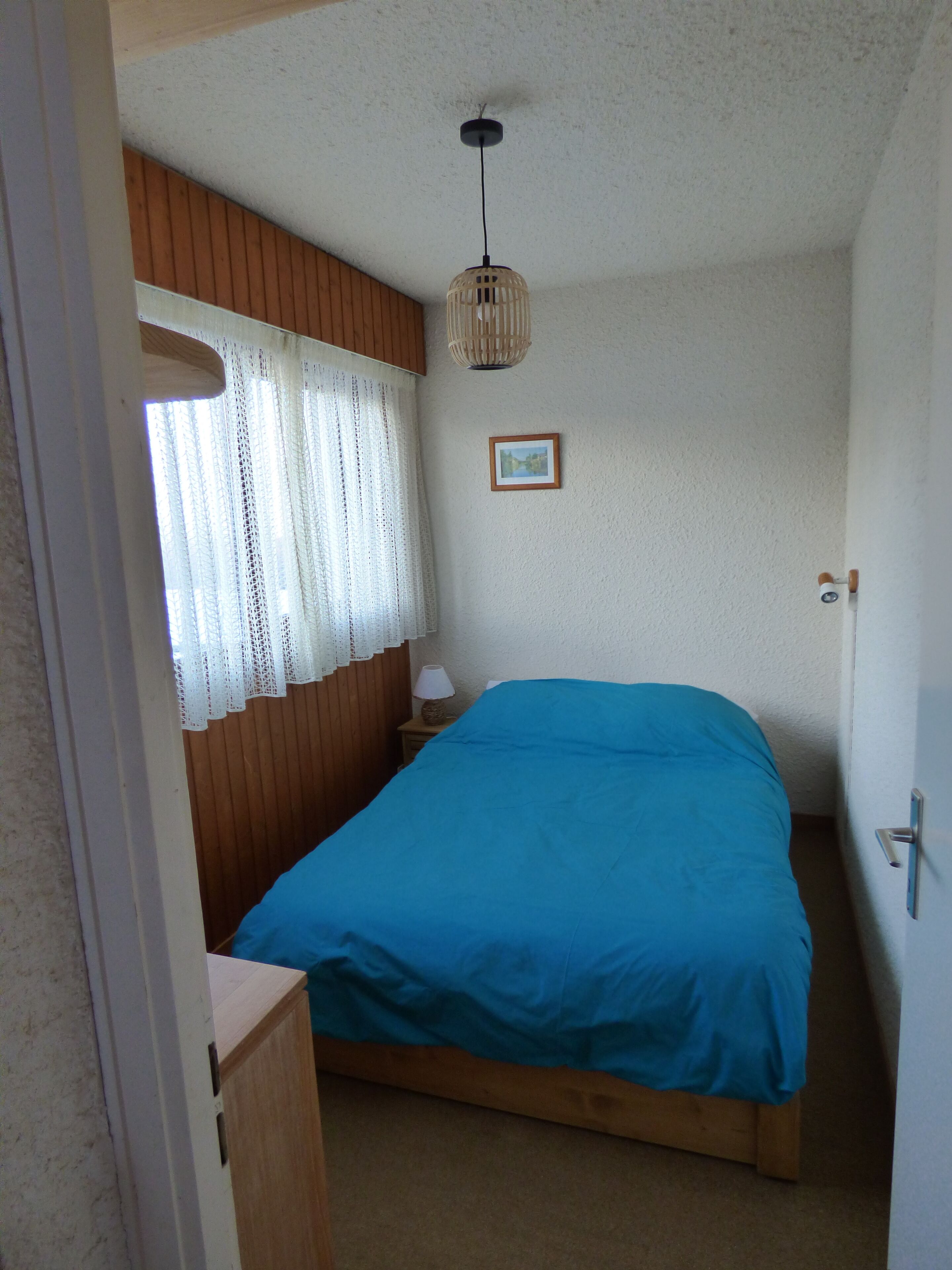 A small, cozy bedroom with a single bed, a wooden headboard, a window with sheer curtains, a hanging light fixture, and a framed picture on the wall.